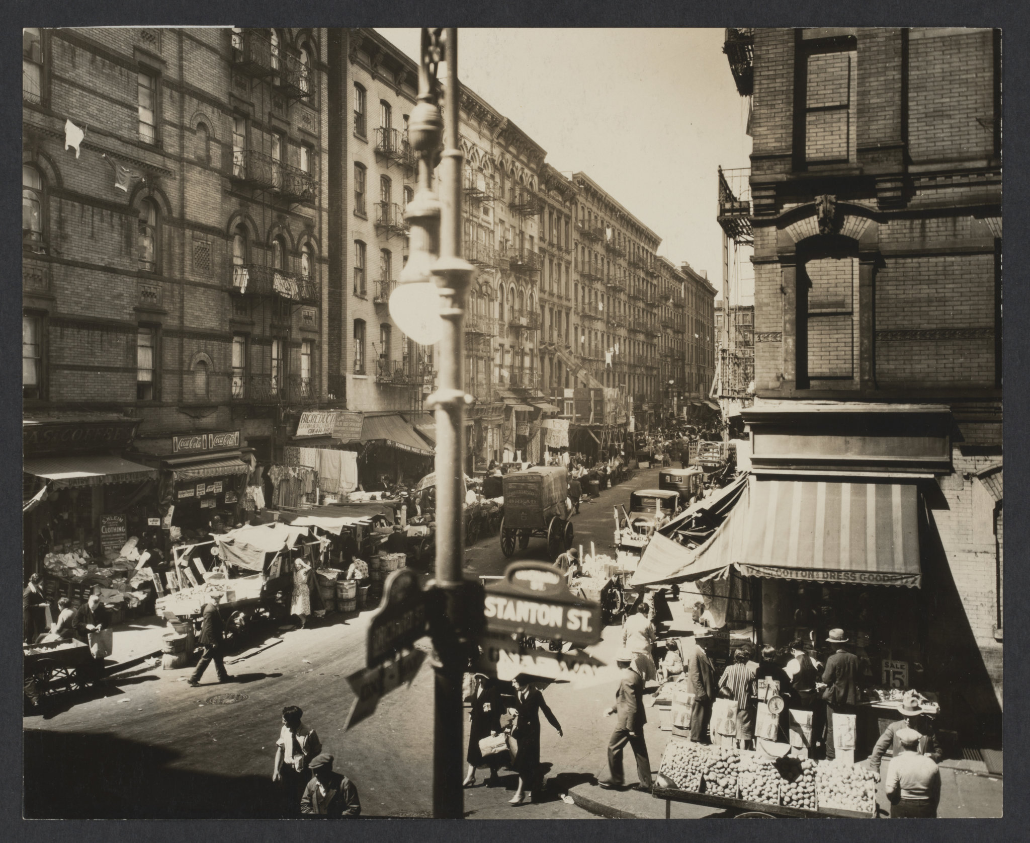 A New York street-scene taken as the camera faces diagonally down the Orchard Street marketplace. A streetlight with attached street signs for Stanton and Orchard is center in the extreme foreground. | Museum of the City of New York. Gift of Joy of Giving Something, Inc., Courtesy of Berenice Abbott